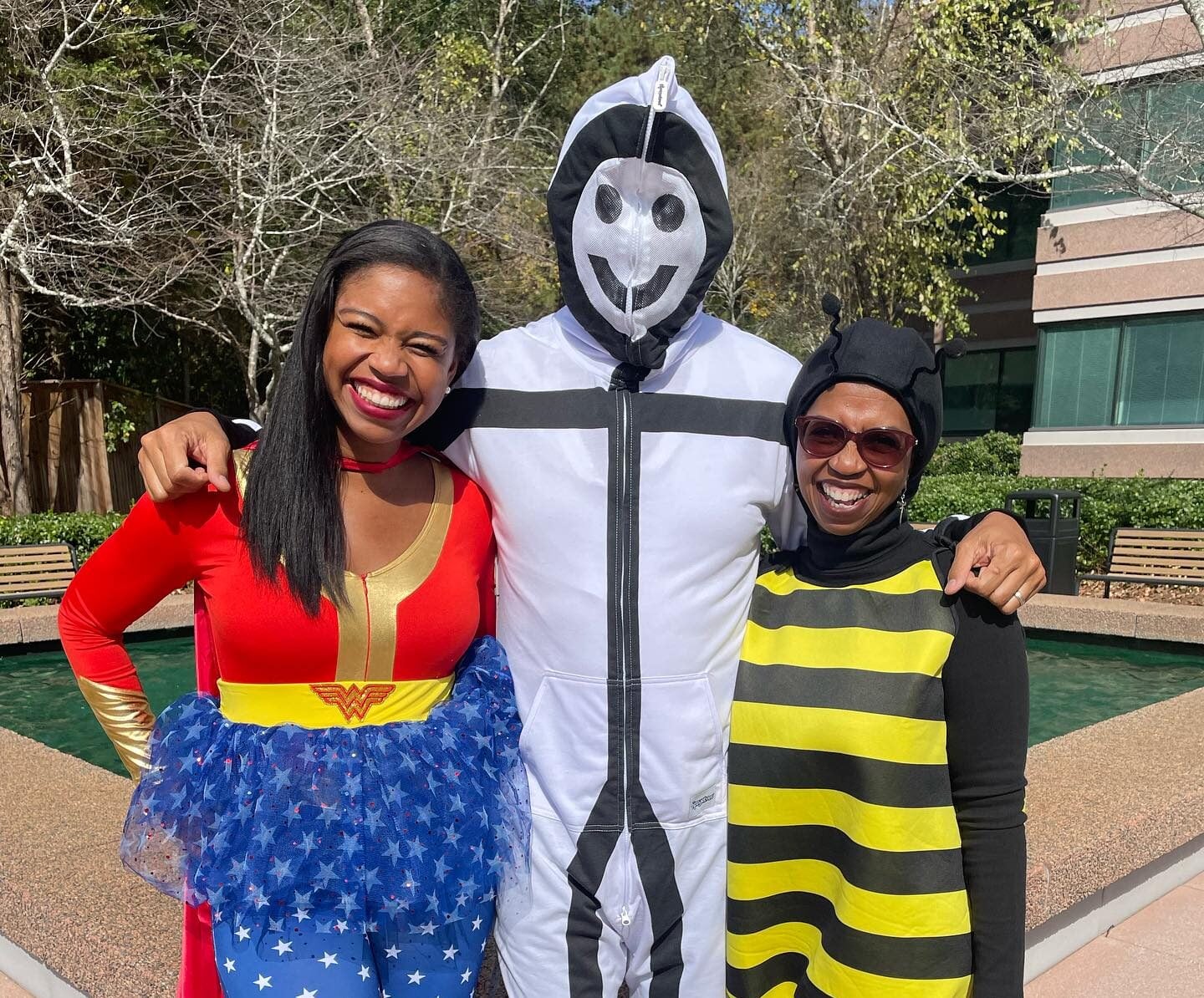Three persons in superhero and bee costumes stand smiling outdoors, with trees and a building in the background.