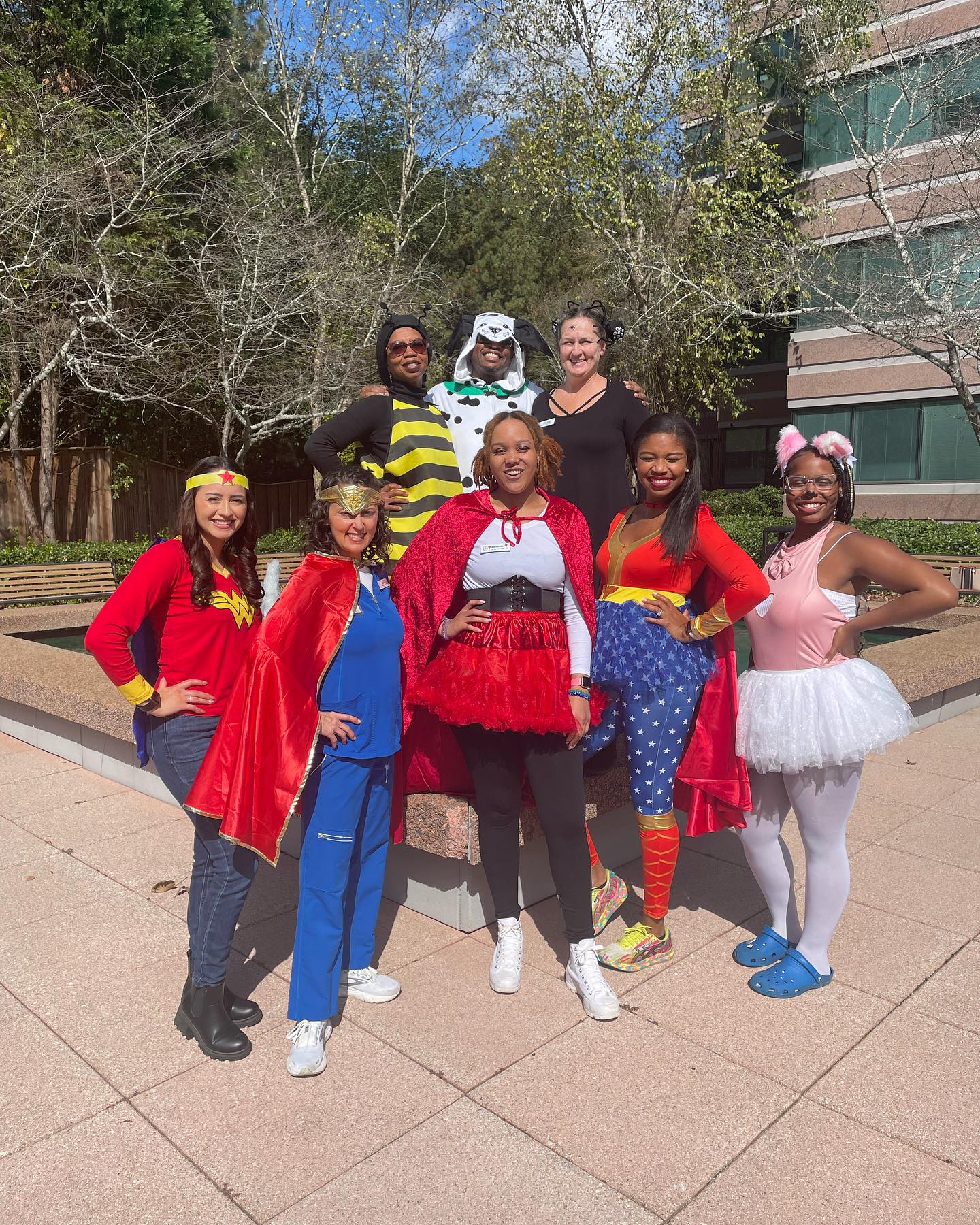 A group of eight costumed people pose outdoors on a sunny day. Trees and a building are in the background.