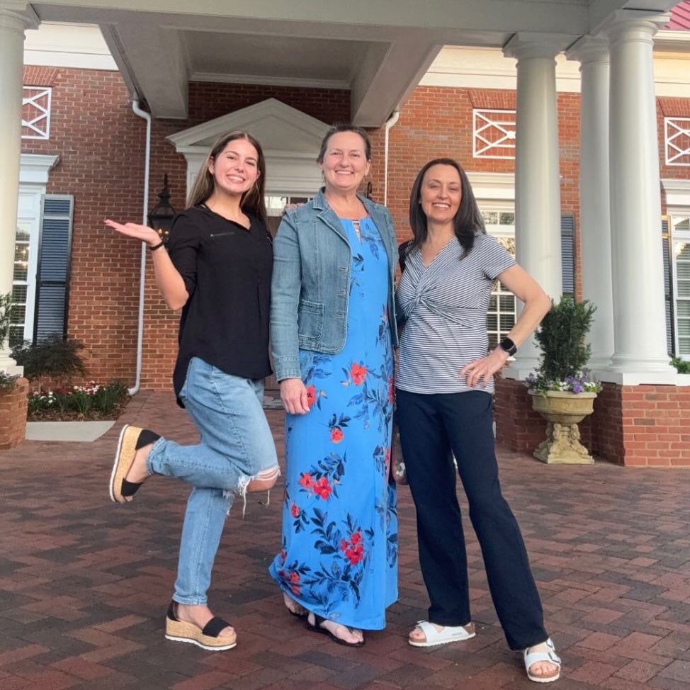 Three people smiling and posing outside a red brick building with white columns and decorative white trim.
