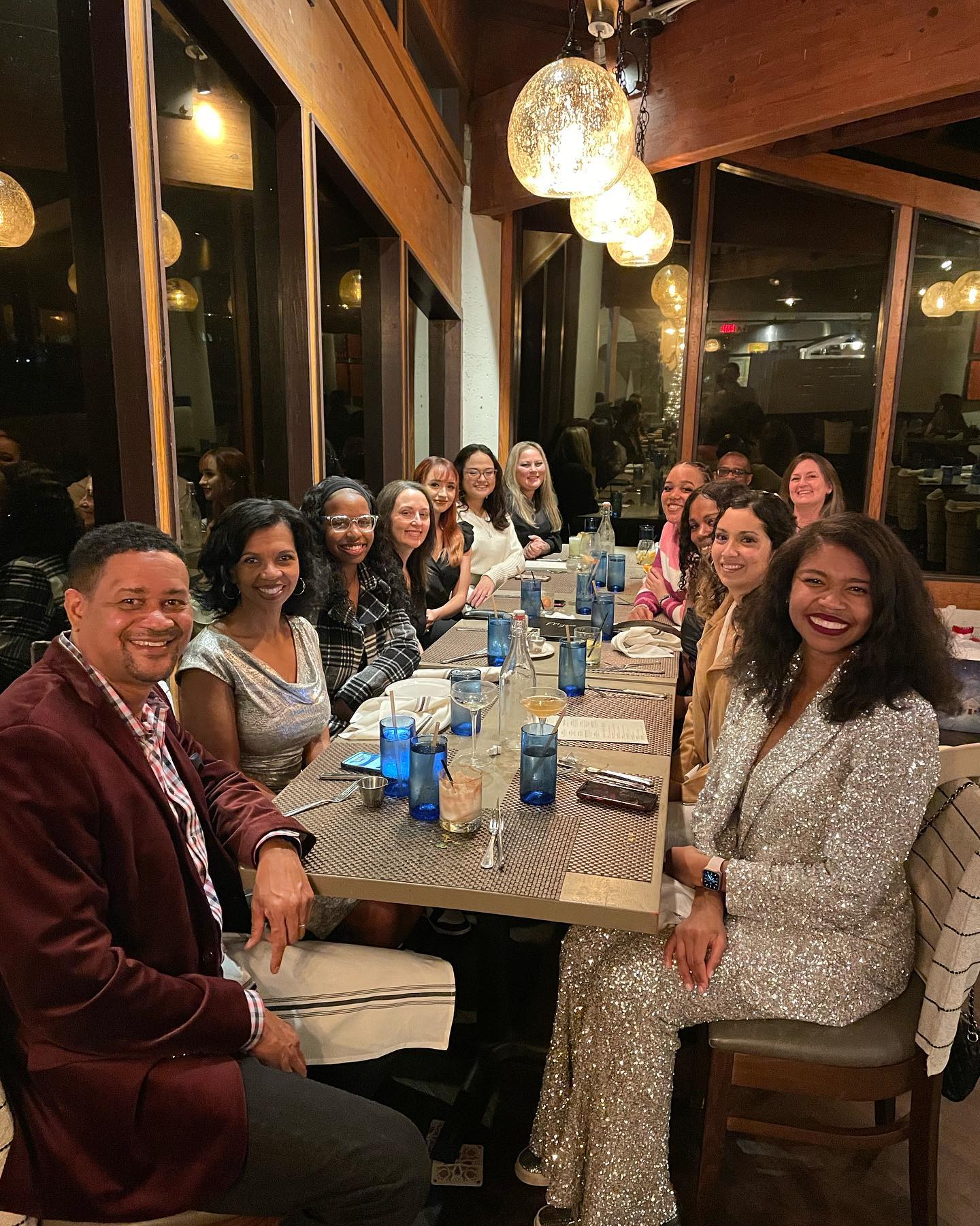 A large group of people enjoys a dinner gathering at a restaurant with a modern interior, featuring hanging lights and large windows.