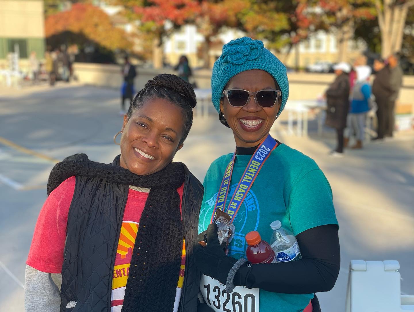 Two persons smile at an outdoor event. One wears a race medal and numbered bib. Autumn foliage and people are visible in the background.